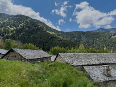 Tepenin yamacında birkaç evi olan bir dağ sırası. Evler taştan yapılmış ve eğimli çatıları var. Gökyüzü açık ve mavi, çimenler gür ve yeşil. Sahne huzur veriyor.