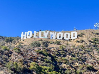 Hollywood tabelası bir tepede. Tepe yeşil ve kayalıktır. Gökyüzü mavi