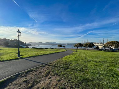 Suya giden yolu olan bir park. Gökyüzü mavi ve bulutlar var. Çimler yeşil, ağaçlar arka planda.