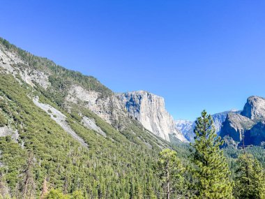 Tepe yüzlü ve arkasında mavi bir gökyüzü olan bir dağ. Dağ ağaçlarla kaplı ve gökyüzü berrak ve parlak.