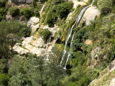 Bir şelale kayalık bir uçurumdan aşağı akıyor. Su yeşil ve temiz. Ağaçlar yemyeşil, kayalar pürüzlü ve pürüzlü. Sahne huzurlu ve sakin, suyun sesiyle.