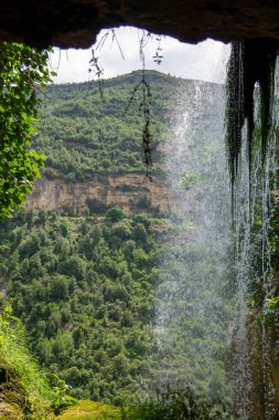 Bir kaya duvarındaki küçük bir delikten bir şelale görülür. Su kayadan fışkırıyor ve çevresi yemyeşil ve yemyeşil.