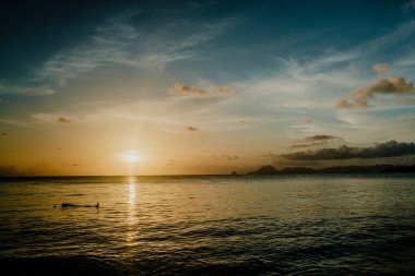 Martinique 'deki Karayip Denizi' nin üzerinde sakin bir günbatımı. Güneş, suya ve renkli gökyüzüne karşı gölgelenmiş uzak dağlara altın yansıyor..