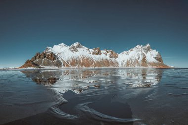 Karla kaplı Vestrahorn dağları güneydoğu İzlanda 'daki Stokksnes plajındaki buzlu sahili yansıtıyor.