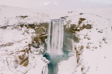 Kış boyunca İzlanda 'daki Skogafoss şelalesinin hava manzarası, üsteki ziyaretçilerle karla kaplı manzaralarla çevrili..