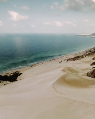 Aerial view of Arher Beach in Socotra, Yemen, showcasing pristine white sand dunes meeting the turquoise waters of the Arabian Sea under a partly cloudy sky. clipart