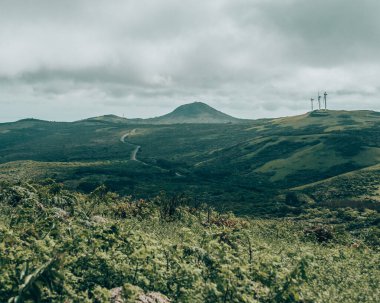 Galapagos, Ekvador 'daki San Cristobal Adası' nın manzarasında bulutlu bir gökyüzü altında yeşil yuvarlanan tepeler ve rüzgar türbinleri yer alıyor.