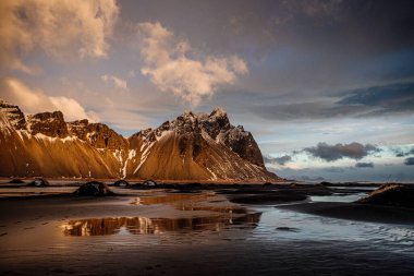 Vestrahorn dağını aydınlatan altın saat ışığı ve İzlanda 'nın güneydoğusundaki siyah kumsal plajını yansıtıyor.