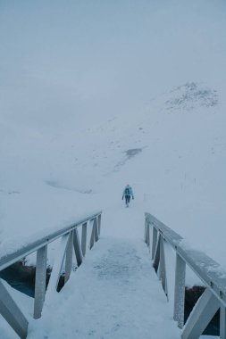 Güney İzlanda, Reykjadalur patikasından karla kaplı bir köprüden geçen bir yürüyüşçü.