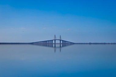 The Sunshine Skyway Bridge gracefully stretches across the calm, reflective Tampa Bay blending seamlessly with the blue sky. The symmetry of the reflection creates a serene and tranquil atmosphere. clipart