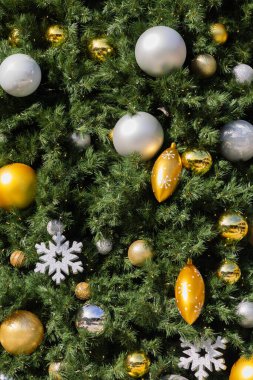 Bir Noel ağacının yemyeşil dalları arasında çeşitli altın ve gümüş mücevherlerin yanı sıra metalik kar tanesi süslemeleri yer alıyor. Dekorasyonlar ışığı yakalıyor, neşeli ve ışıl ışıl bir tatil atmosferi yaratıyor..