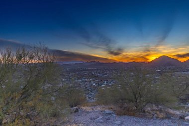 Canlı bir günbatımı, Tucson Arizona üzerinde turuncu ve mavi renkli çarpıcı bir görüntü sergiliyor. Şehir manzarası engin çöl manzarasının ortasına sakince yerleşmiş durumda, saguaro kaktüsü ve diğer çöl bitkileri araziyi benekliyor..