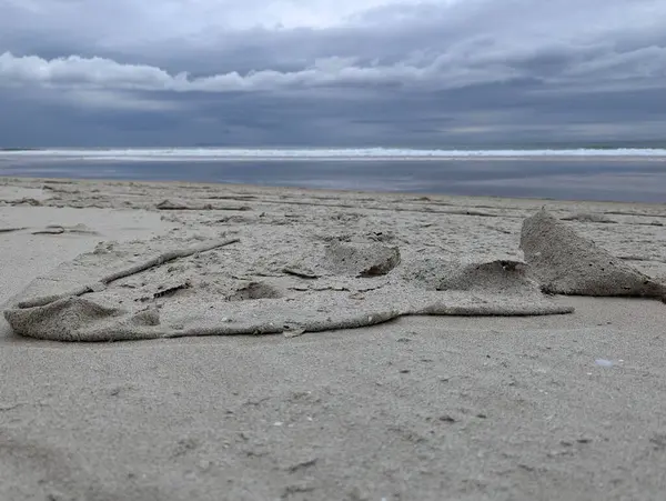 Görüntü, sahilde sakin bir sahne yakalıyor. Ön planda muhtemelen rüzgar ya da gelgit tarafından yaratılmış karmaşık desenli kum bulunur. Okyanus arka planda görülebilir, kıyıya doğru yumuşak dalgalar akıyor..