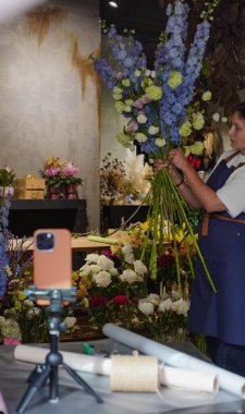 Çiçekçi, internetten buket yapmak için üç ayaklı bir masaya yerleştirilmiş bir telefonu kullanıyor. Çiçekçi eğitici bir video kaydediyor. Küçük bir iş. Yakın plan
