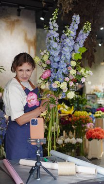Çiçekçi, internetten buket yapmak için üç ayaklı bir masaya yerleştirilmiş bir telefonu kullanıyor. Çiçekçi eğitici bir video kaydediyor. Küçük bir iş. Yakın plan