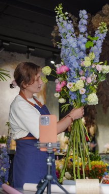 Çiçekçi, internetten buket yapmak için üç ayaklı bir masaya yerleştirilmiş bir telefonu kullanıyor. Çiçekçi eğitici bir video kaydediyor. Küçük bir iş. Yakın plan