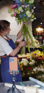 Çiçekçi, internetten buket yapmak için üç ayaklı bir masaya yerleştirilmiş bir telefonu kullanıyor. Çiçekçi eğitici bir video kaydediyor. Küçük bir iş. Yakın plan