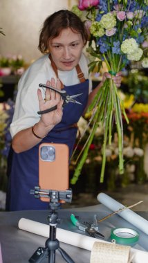 Çiçekçi, internetten buket yapmak için üç ayaklı bir masaya yerleştirilmiş bir telefonu kullanıyor. Çiçekçi eğitici bir video kaydediyor. Küçük bir iş. Yakın plan. Kullandığı budama makinesini gösteriyor..