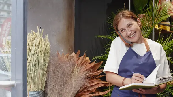 Çiçekçi telefonda konuşuyor, gülümsüyor ve not defterine iş notları yazıyor. Küçük bir iş. Yakın plan.