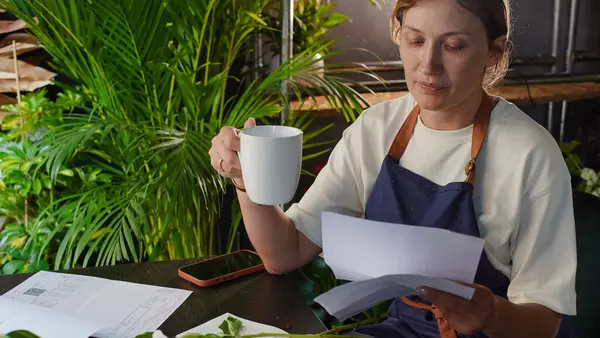 Bir çiçekçinin sahibi bir masada otururken belgeleri gözden geçiriyor ve kahve içiyor. Küçük bir iş. Yakın plan