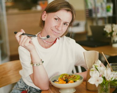 Kız kameraya bakıyor, gülümsüyor, elinde bir kaşık tutuyor ve meyve salatası yemek üzere. Sağlıklı yemek. Bir kafede kahvaltı. Yakın plan. Yumuşak odak.