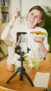 Kız elinde bir tabak tutuyor, gülümsüyor ve telefon kamerasına yemek gösteriyor. Çevrimiçi yemek kursları. Yumuşak odaklanma. Yakın plan.