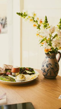 Kafedeki masada kahvaltılı bir tabak var, üzerinde birkaç dilim ekmek, salata, tereyağı, haşlanmış yumurta var. Masanın üzerinde çiçekli bir vazo var. Kahvaltıda. Yakın plan. Yumuşak odak.