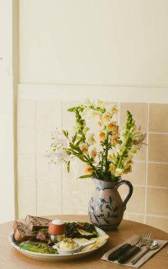 Kafedeki masada kahvaltılı bir tabak var, üzerinde birkaç dilim ekmek, salata, tereyağı, haşlanmış yumurta var. Masanın üzerinde çiçekli bir vazo var. Kahvaltıda. Yakın plan. Yumuşak odak.