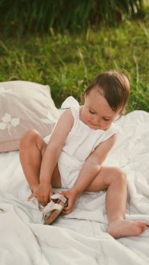Beyaz elbiseli küçük kız parkta battaniyede oturan sandaletleri giymeyi öğreniyor. Yumuşak odaklanma. Yakın plan
