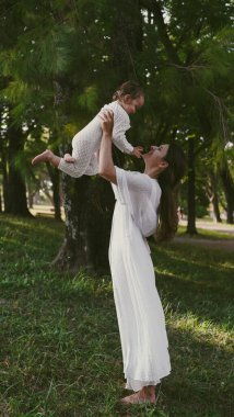 Beyaz elbiseli genç bir anne parkta çimlerin üzerinde dururken küçük kızını onun üzerine atar. Yumuşak odak