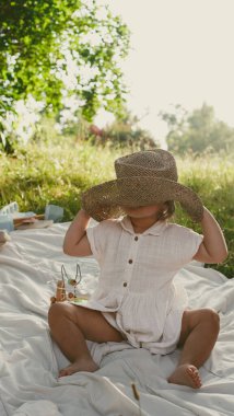 Küçük kız parkta battaniyenin üzerinde otururken büyük bir hasır şapkayla oynuyor. Yumuşak odaklanma. Yakın plan