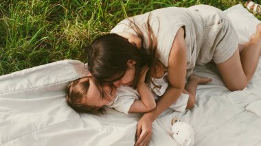 Anne ve küçük kızı parkta battaniyeye uzanmış sarılıp gülüyorlar. Yumuşak odaklanma. Yakın plan