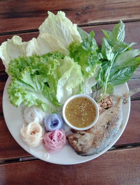 Geleneksel Tayland salatası malzemeleri ve ızgara uskumru ile ahşap bir masada sunulan bir tabak sosun yanında.