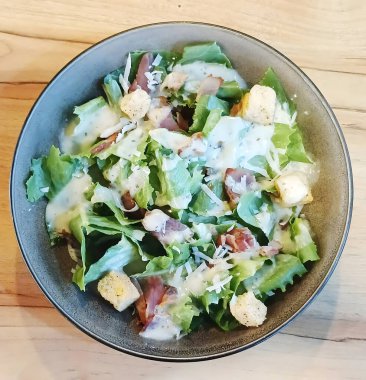 Bir kase Sezar salatası, kızarmış ekmek ve kremalı Sezar soslu parmesan peyniri. Salata ahşap bir masanın üzerinde..