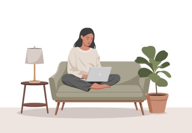 Kadın, gümüş bir dizüstü bilgisayar kullanarak yeşil bir koltukta bağdaş kurmuş oturuyor. Terracotta saksı bitkisi ve masa lambası yakınlarda..