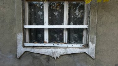 Eski bir pencere, yıllar süren terk edilmeyi yansıtan ihmal edilmiş dış ve dağınık yapraklarla çevrili, ince dantelli bir perde ve boyayı içerir..