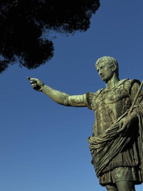 Bir Roma figürünün heybetli heykeli dimdik ayakta duruyor ve bir kolu uzatılmış şekilde öne doğru hareket ediyor. Şekil geleneksel zırhlarla süslenmiş, yukarıdaki canlı mavi gökyüzünü tamamlıyor..