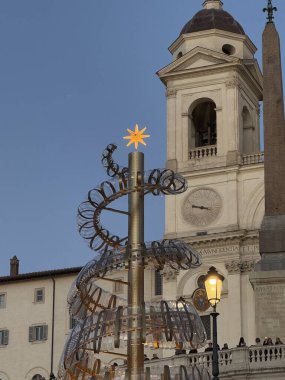 Şenlikli bir bayram dekorasyonunda, üzerinde parlayan bir yıldız bulunan sarmal bir tasarım, eski bir kilisenin arka planına ve akşam çökerken bir dikilitaşa dayanır..
