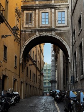 Ziyaretçiler Roma 'da dar bir sokakta yürüyor. Tarihi bir kemer iki binayı birbirine bağlıyor. Motosikletler kaldırım taşı yolu boyunca park halindedir. Tam bir İtalyan atmosferi yaratırlar..