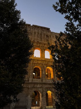Güneş batarken, Roma 'daki antik Kolezyum akşam gökyüzünün altında parlıyor. Koyu yeşillikle çerçevelenmiş, mimari ihtişamını ve zengin tarihini gözler önüne seriyor..