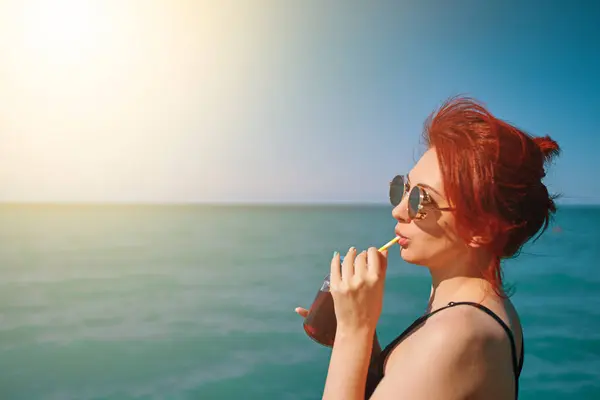 Girl on summer vacation. Red-haired woman in sunglasses drinks lemonade from bottle through cocktail tube. Blue sea, sunlight and clear sky on background. Copy space for travel agency advertising.