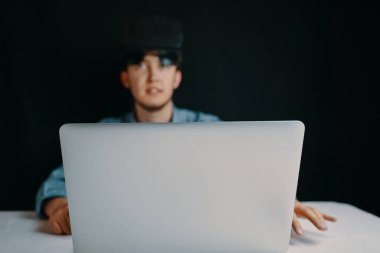 VR kulaklıklı genç bir adam loş bir odada dizüstü bilgisayar kullanmaya odaklanır ve yüksek teknoloji nişanını gözler önüne serer.