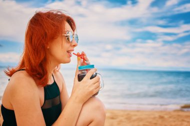 Mayo giymiş sakin bir bayan sahilde kokteyl içip yüzünü çeviriyor. Tropik alkol içeceği. Bulutlu bir yaz günü. Tatilde güneş gözlüklü kızıl saçlı kadın. Denizde tatiller.