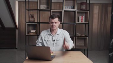 İnsan kameraya bakar ve elleriyle hareket ederken konuşur. Gömlekli ciddi bir adam bir masada dizüstü bilgisayarla oturup röportaj yapıyor. Arkaplanda kitaplar olan raflar.