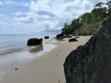 Önünde büyük bir kaya olan kayalık bir sahil. Kayalar sahile saçılmış ve Nibung Indah sahilinde su sakin.