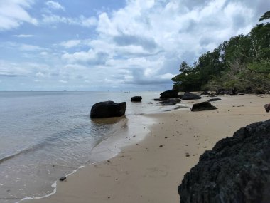 Önünde büyük bir kaya olan kayalık bir sahil. Kayalar sahile saçılmış ve Nibung Indah sahilinde su sakin.