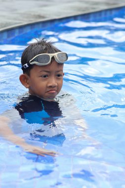Asyalı küçük çocuk, güneşli bir havuzun kenarında neşe dolu bir an yaşarken kameraya doğru neşeli yüzler yapıyor..