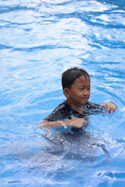 Asyalı küçük çocuk, güneşli bir havuzun kenarında neşe dolu bir an yaşarken kameraya doğru neşeli yüzler yapıyor..