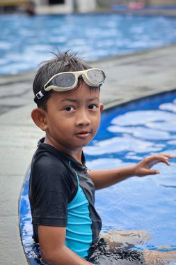 Asyalı küçük çocuk, güneşli bir havuzun kenarında neşe dolu bir an yaşarken kameraya doğru neşeli yüzler yapıyor..
