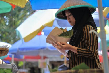 Balikpapan-Endonezya 29 Mayıs 2025 Geleneksel Cava kıyafetleri ve pelerinli kız (Cava şapkası) Geleneksel bir pazarda müşterilere hizmet veriyor